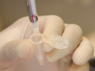 A pipette with fluid sits inside a sample receptacle held by a gloved hand.