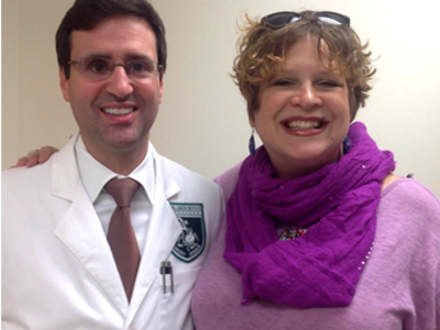 Dr. Christian Hinrichs and Susan Scott stand looking at the camera and smile. Dr. Hinrichs has short dark hair, is wearing glasses, a white coat, and shirt and tie. Susan has short, dirty blonde hair, is wearing sunglasses on top of her head and a large purple scarf and top.