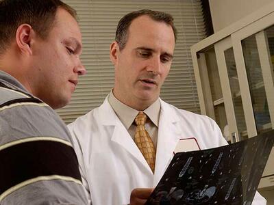A male doctor holds an x-ray he is looking at while speaking to a male patient. The doctor has short dark hair and wears a white lab coat with shirt and tie. The patient has a yellow-and-black polo shirt. Window blinds and a cabinet are in the background.