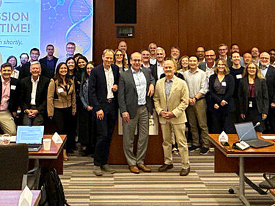 A large group of workshop attendees poses for a photo in a large meeting room. A screen behind them reads, “It’s session break time! We will return shortly.”