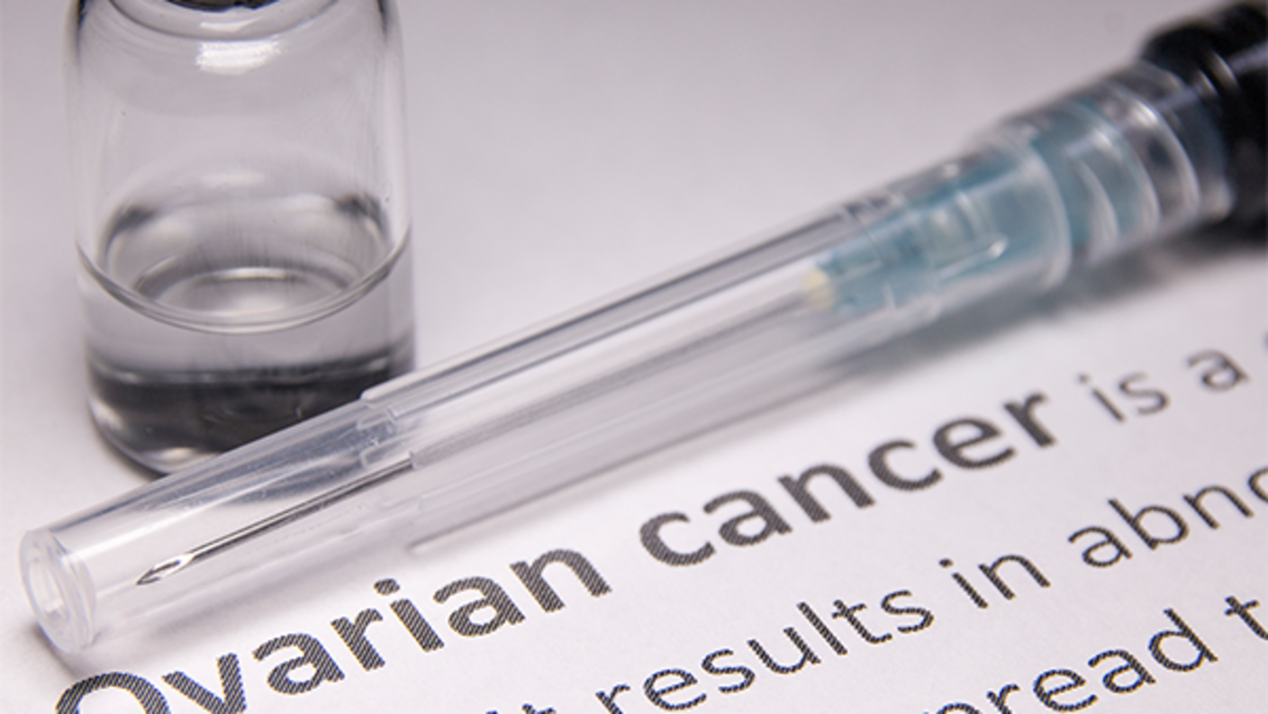 A close-up of a syringe and medical bottle containing clear liquid. These two items are sitting on a piece of paper that shows the definition of ovarian cancer. The words “ovarian cancer” are bold; the rest of the text is mostly cropped off the image, though what is shown indicates it is a definition of ovarian cancer.