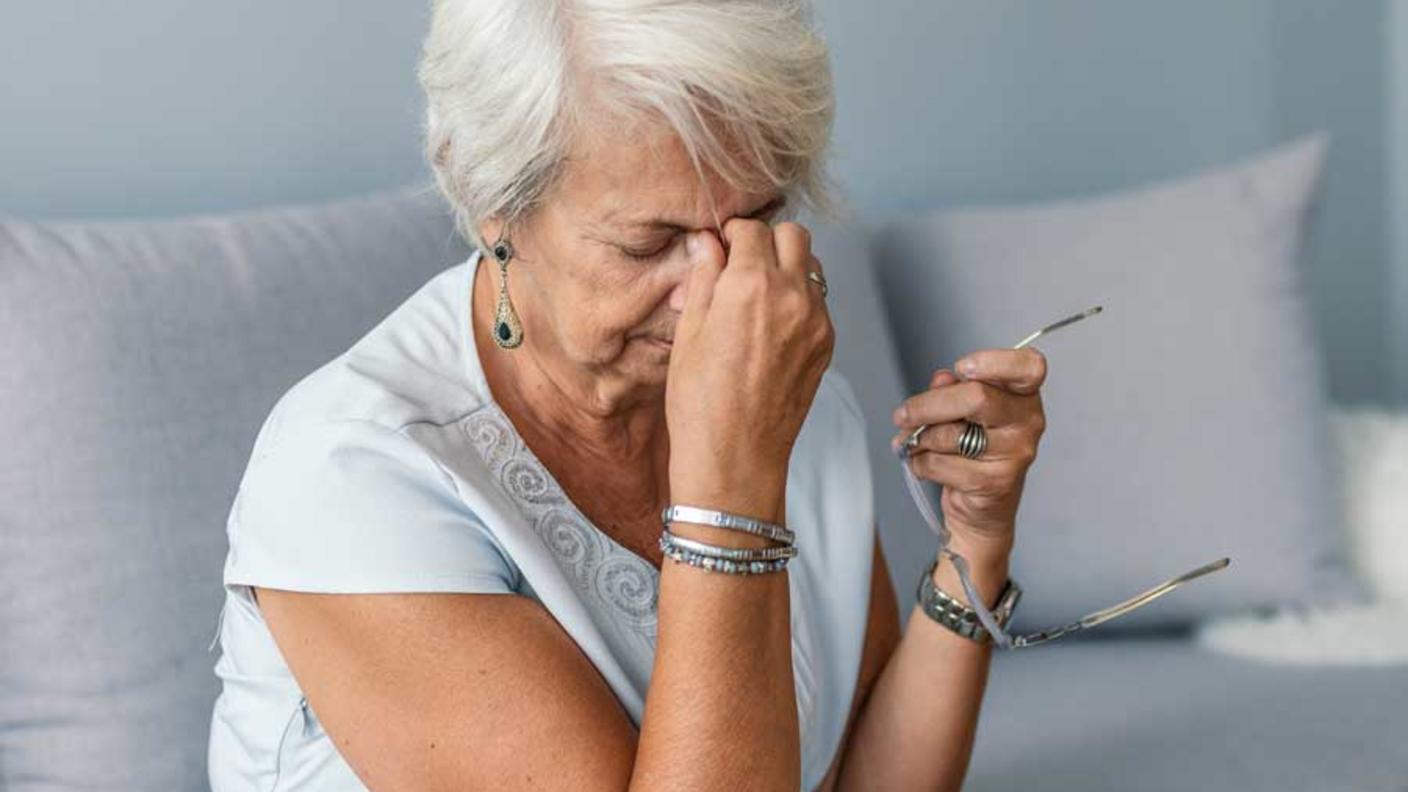 An elderly woman sits on a couch holding her glasses in one hand and rubbing her forehead with the other, appearing to be in discomfort.
