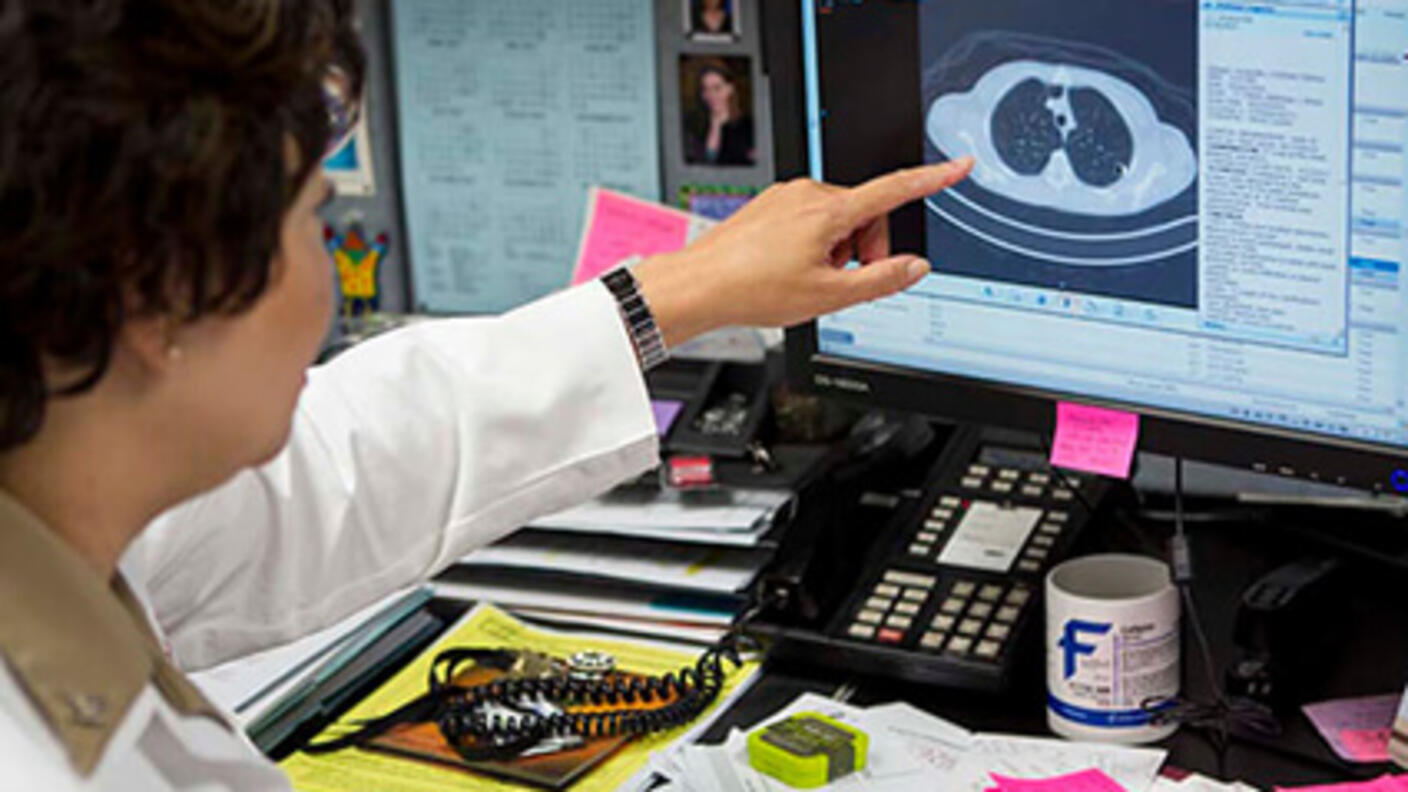 Researcher seated by a desk and computer screen points to detail in a CT scan on the screen.