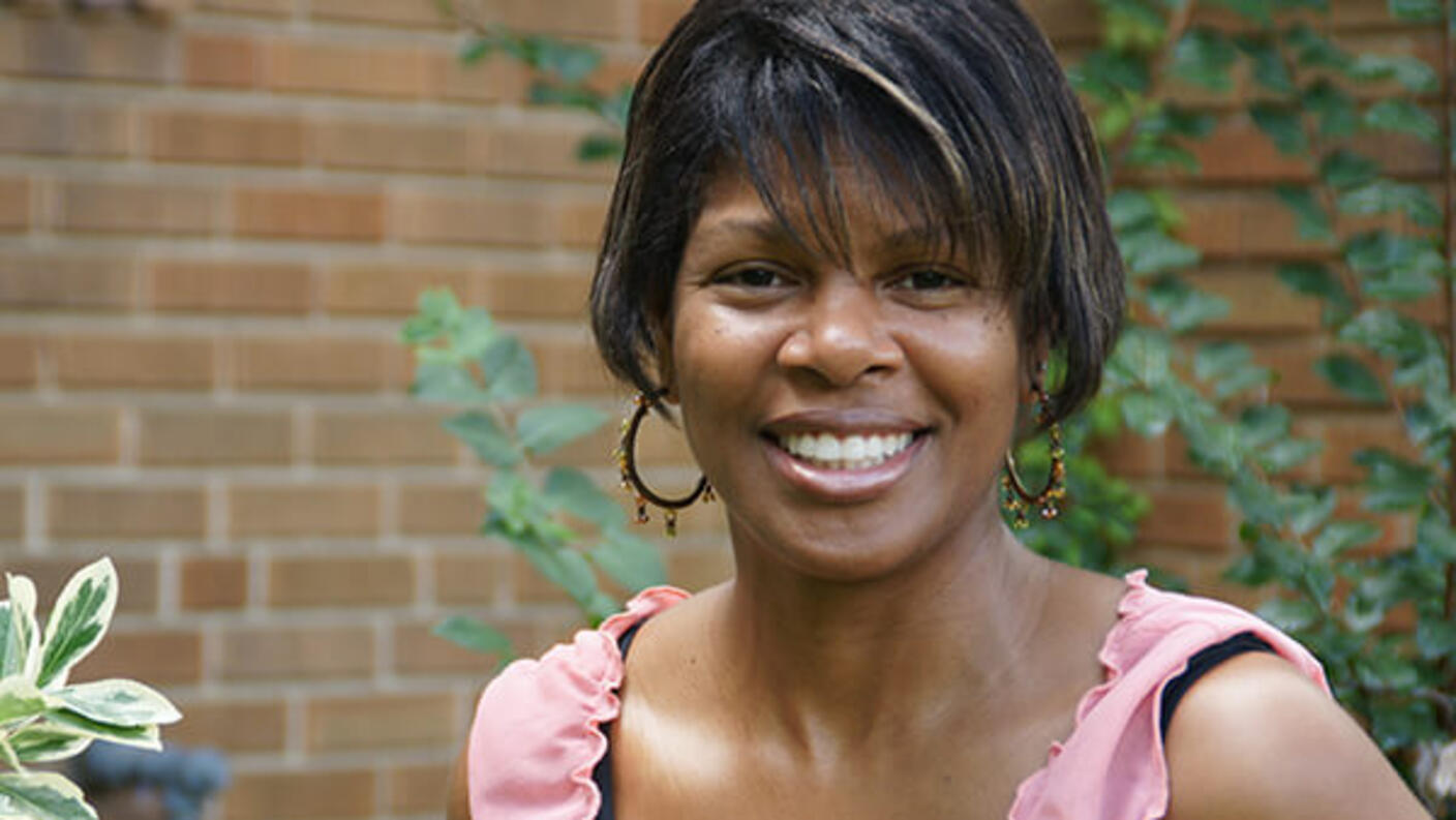 Portrait of Melanie Nix, smiling while wearing a pink ruffled top and earrings, photographed outdoors with greenery and brick in the background