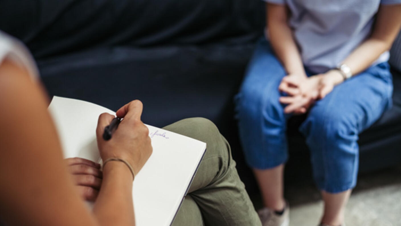 Therapist taking notes during a session with a cancer patient, shown seated with hands clasped