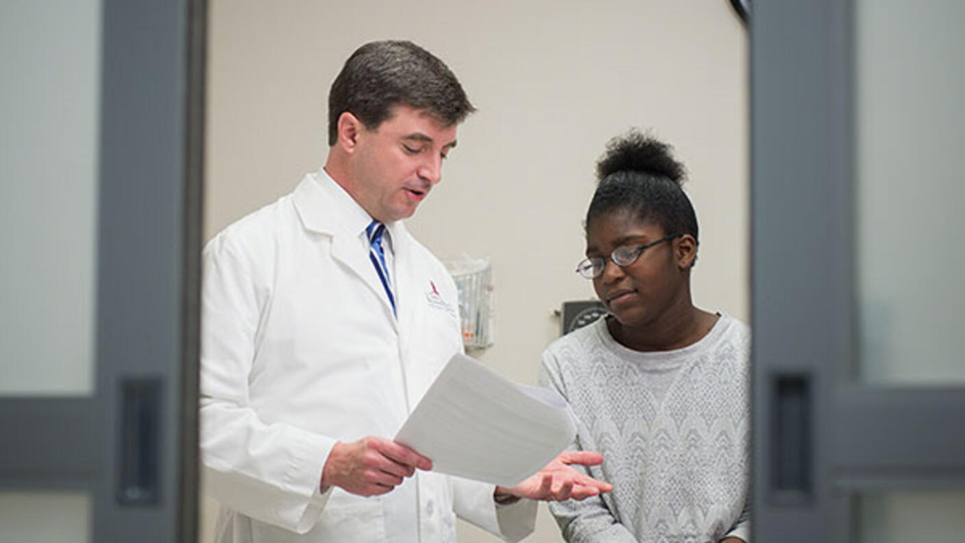 A doctor looks at a piece of paper and speaks to his patient. The doctor is a white middle-aged male, wearing a lab coat, button up shirt and tie, He is standing and gesturing as he speaks. The patient, a younger Black woman with glasses, a long sleeve grey sweater and jeans, sits on the exam table and looks slightly distressed. Out of focus doors to the room are in the foreground.