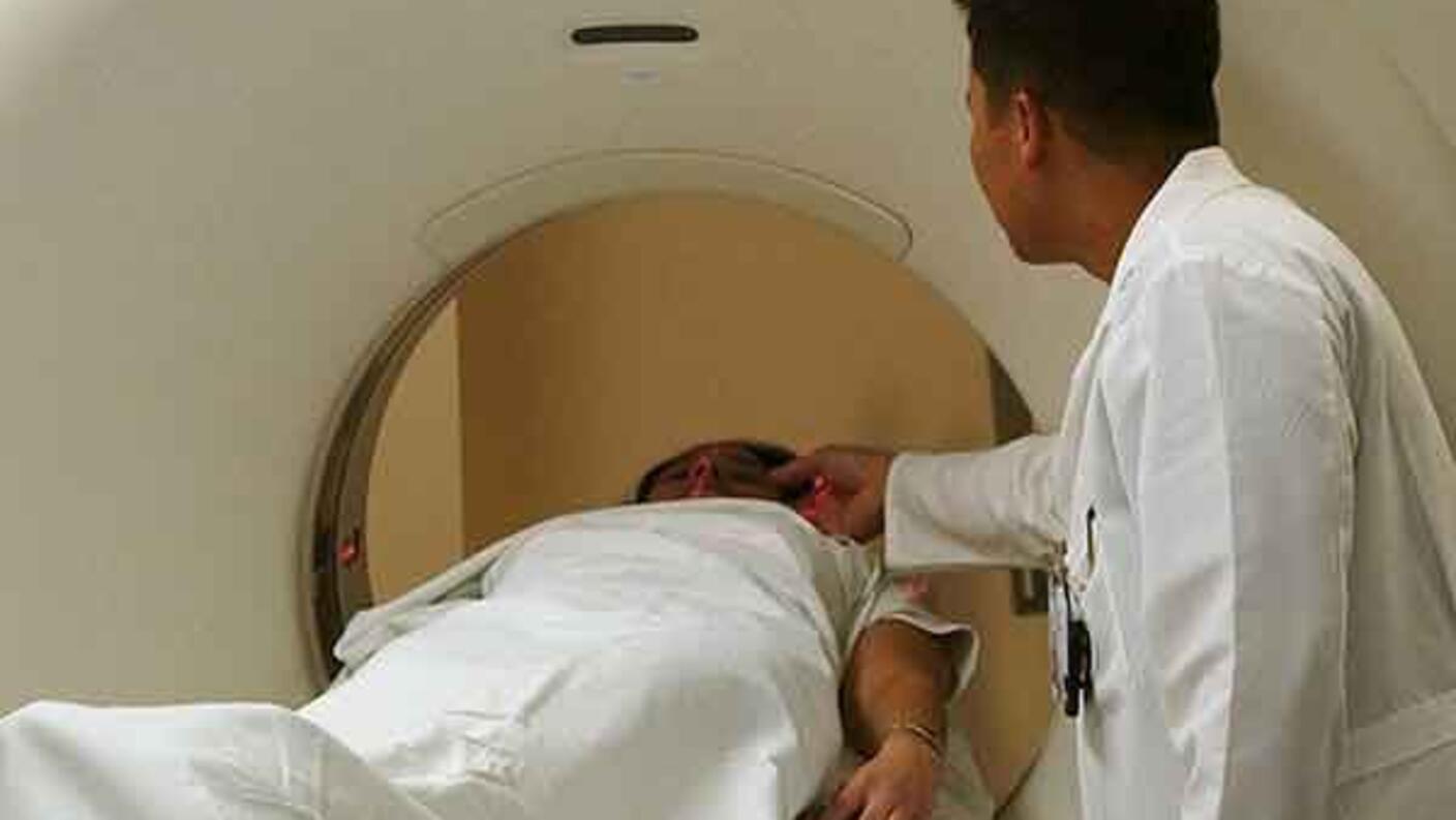 A doctor in a white coat watches as a patient covered in a white blanket goes into a CT machine. The doctor’s hand is partially in the machine.