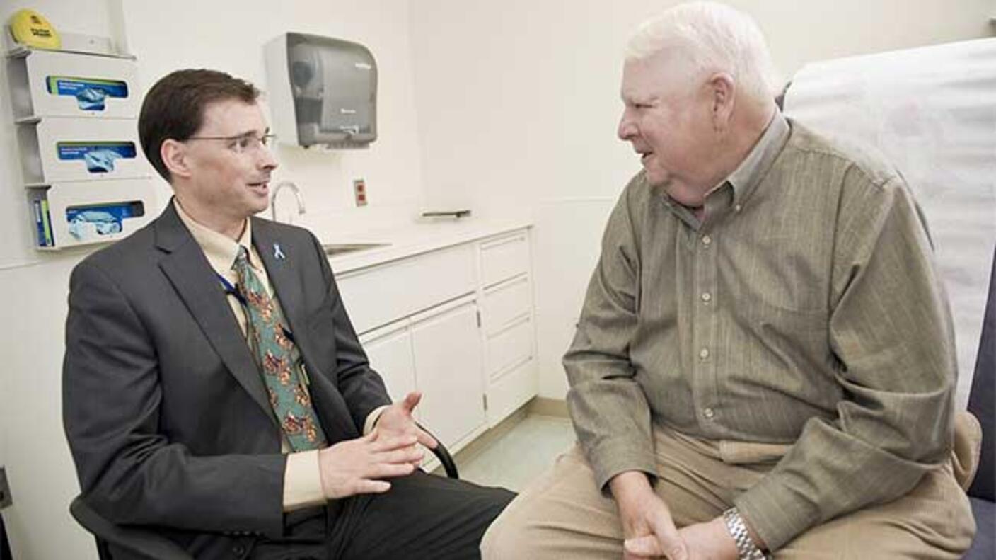 A doctor and a patient, both sitting, speak to each other. The doctor is middle aged with short dark hair, glasses, a grey suit and tan shirt with patterned tie. He’s sitting in a chair. The patient is an elderly man in a green button up shirt and khakis with white hair; he sits on an exam chair. A sink, cabinets, and gloves are in the background.