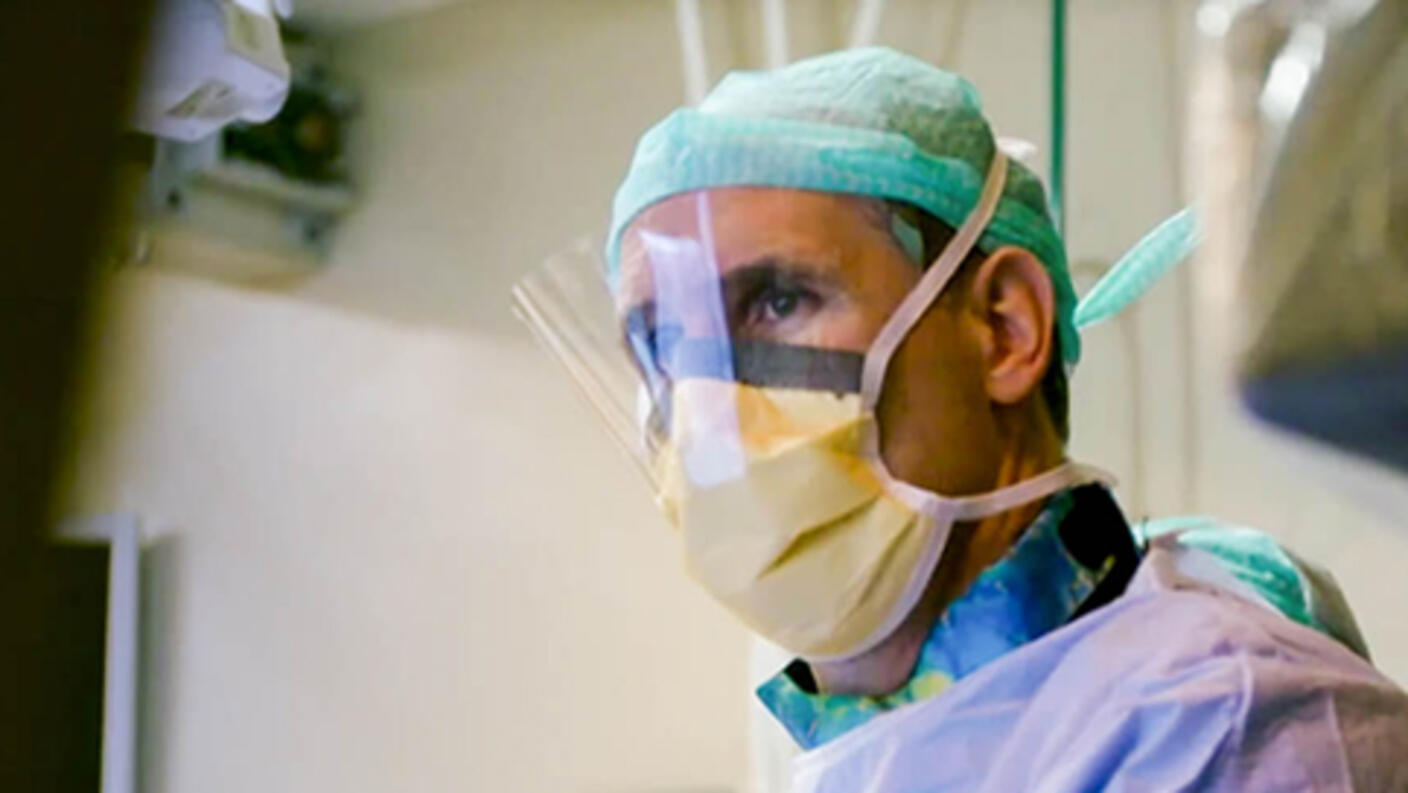 A doctor in an operating room wears a plastic face shield, a mask, scrubs and a green cap. He looks off into the distance. In the bottom left corner, text reads “F1RST in HUMAN” with the dates and times of August 10, 17 & 24, 9pm ET/PT. The Discovery Channel logo is on the bottom right corner.