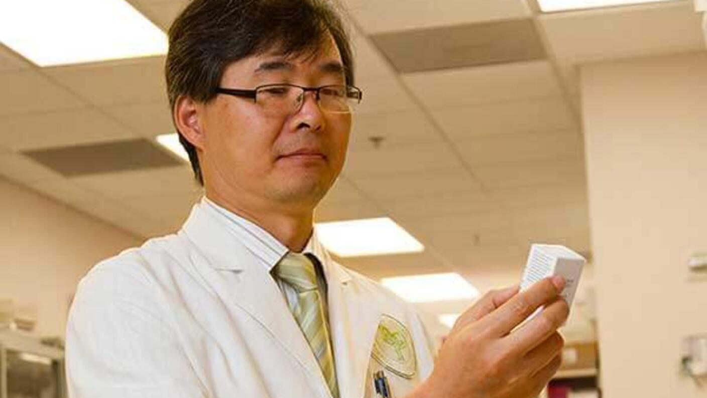 A pharmacist with short black hair and glasses stands in a pharmacy examining a boxed drug.