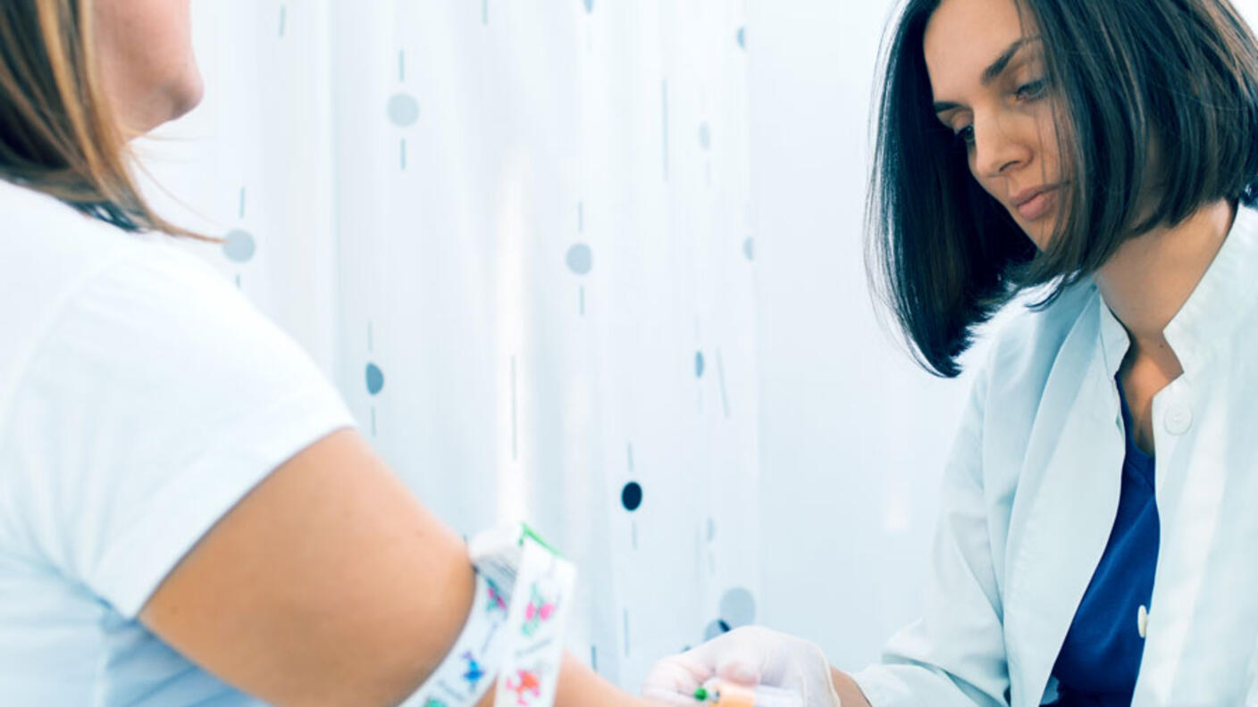 Healthcare professional in white coat drawing blood from a patient's arm. The patient wears a white shirt and has a colorful tourniquet around their upper arm