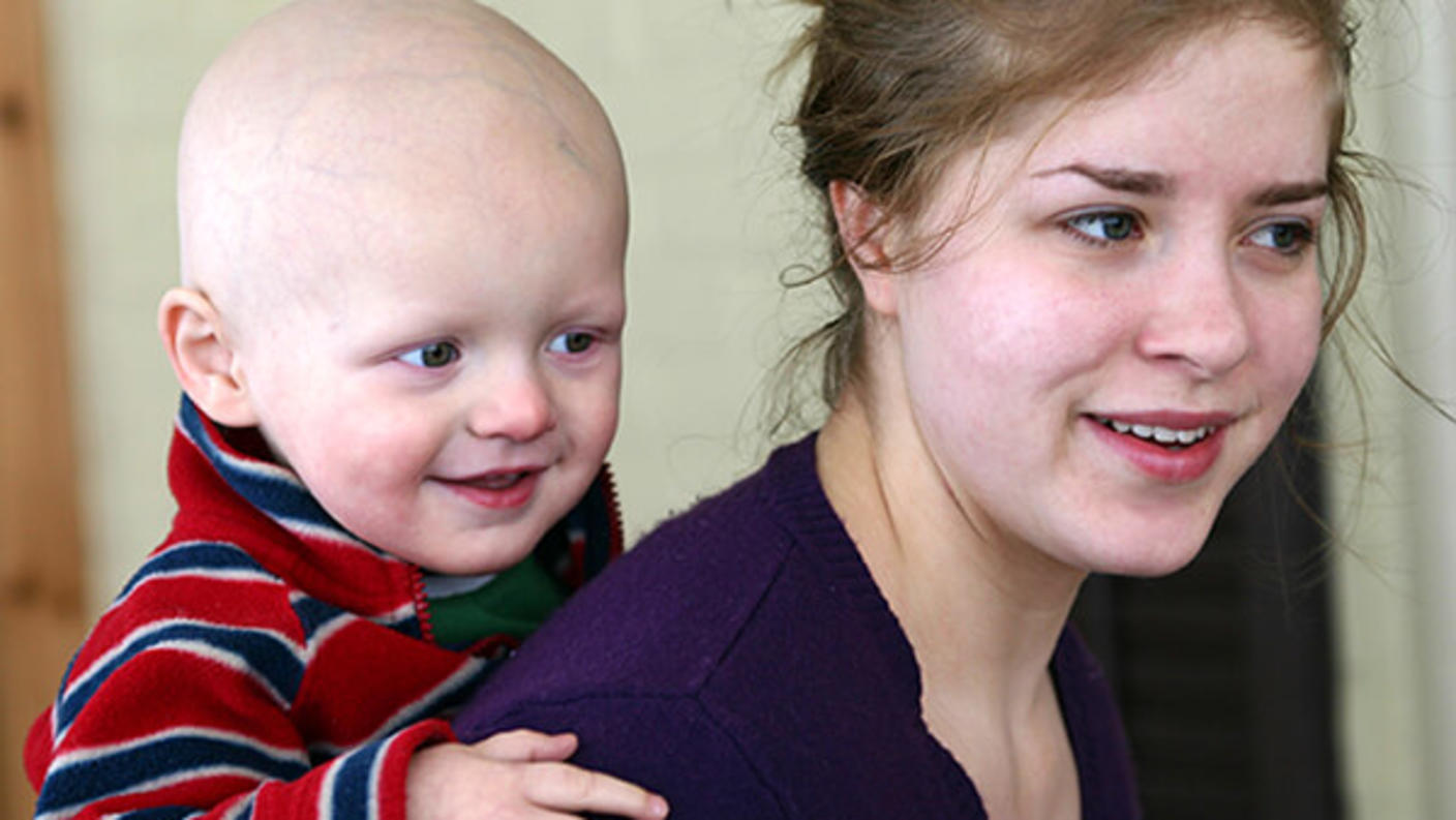 Child with a bald head, is smiling while playfully holding onto the woman’s shoulders. The woman looks content as she interacts with the child.
