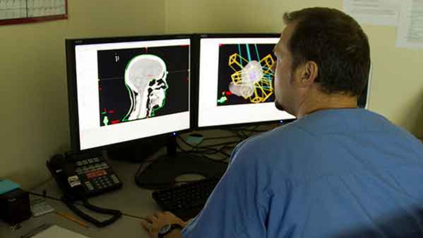 Medical professional in scrubs reviewing brain scan images on dual computer monitors, showing both traditional X-ray and enhanced colorized views.