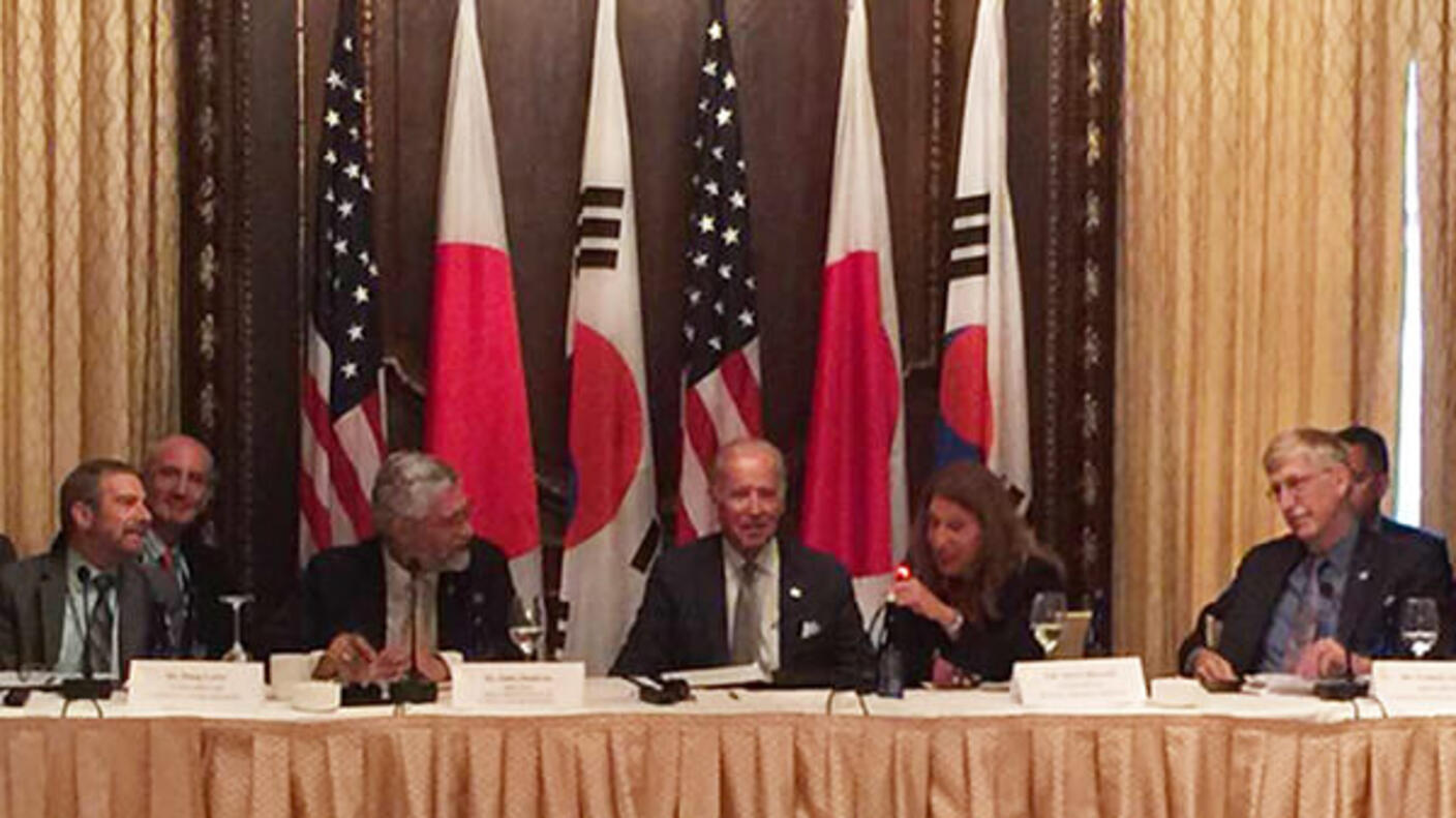 Political and health leaders sit at table with the flags of USA, Japan, and Korea behind them