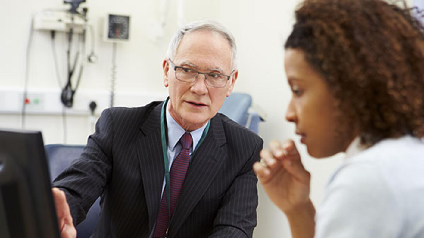 A doctor points at a computer screen while speaking to a patient. The doctor is an elderly man with white hair, glasses, and a dark suit with a lanyard around his neck. He is looking at the computer screen, not the patient. The patient is an adult woman with curly brown hair and a long light blue shirt.