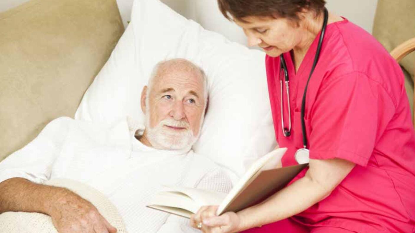 Nurse in pink scrubs reading to an elderly male patient in bed as part of palliative care, focusing on comfort and quality of life during cancer treatment.