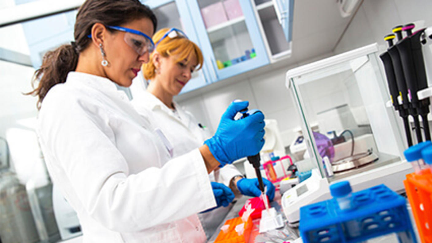 Two female scientists stand at a laboratory bench and use pipettes to prepare samples.