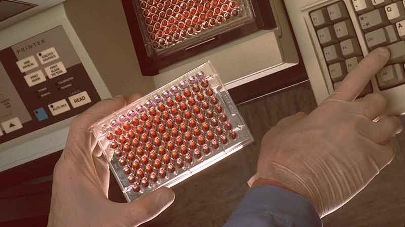Scientist holding a tray of cell lines in their left hand and pressing buttons on a computer keyboard with their right hand.