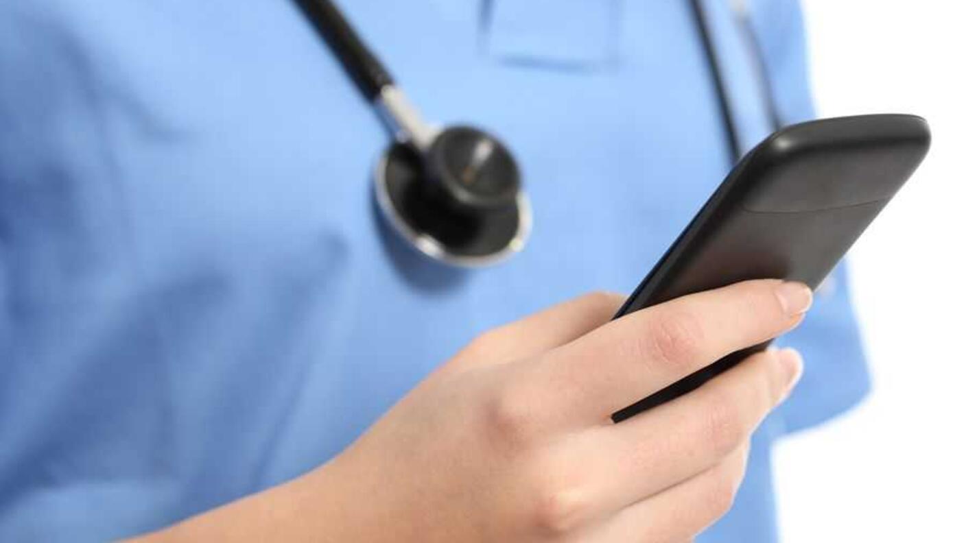 A close-up of hand holding a cell phone. The person’s body, wearing blue scrubs and a stethoscope around their neck, is blurry yet visible in the background.