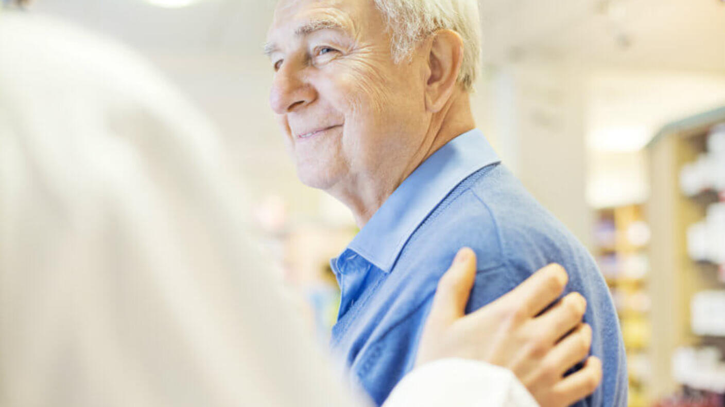 Smiling elderly man in blue shirt receiving a supportive hand on his shoulder from someone off-camera 