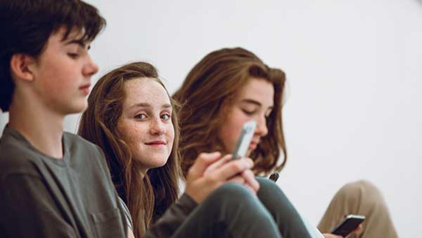 Three teenagers sitting together, with the girl in the middle smiling at the camera while her friends look at their phones in a casual, relaxed setting.