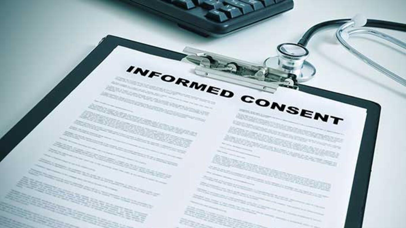 A close-up of a hand holding a pen to a clipboard holding a piece of paper. The words “Informed Consent”  are printed in large letters at the top of the paper, with smaller text in two columns below. A stethoscope and part of a keyboard are in the background.
