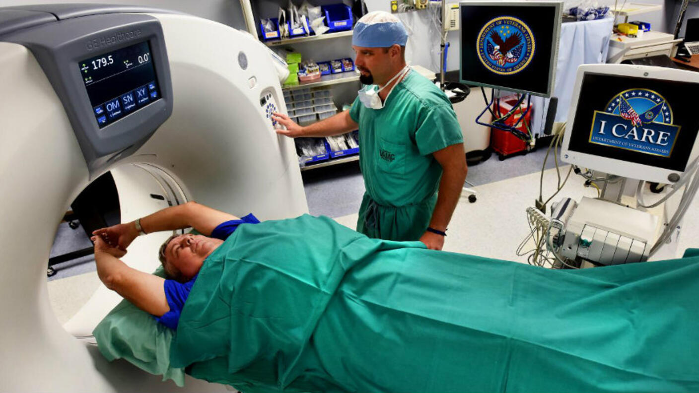 Patient lying on CT scan table receiving low-dose screening for lung cancer, with medical technician operating the machine in a hospital setting displaying Veterans Affairs and ICARE logos on equipment monitors.