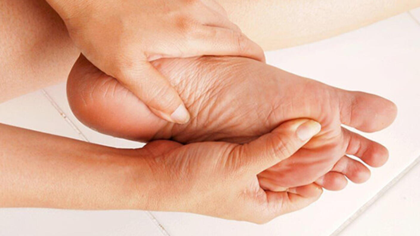 A close-up of a woman’s hands holding her foot. Her left hand ism mostly around the area of her heel; the right hand rubs the bottom of the foot below her big toe. A tile floor can be seen in the background.