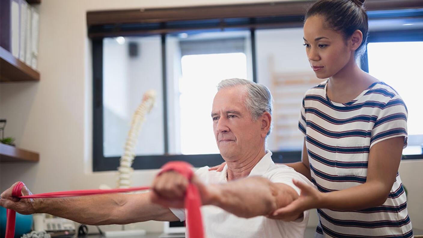 Physical therapist assisting an older male patient with resistance band exercises in a bright therapy clinic, with the patient stretching the red resistance band while the therapist provides guidance and support.