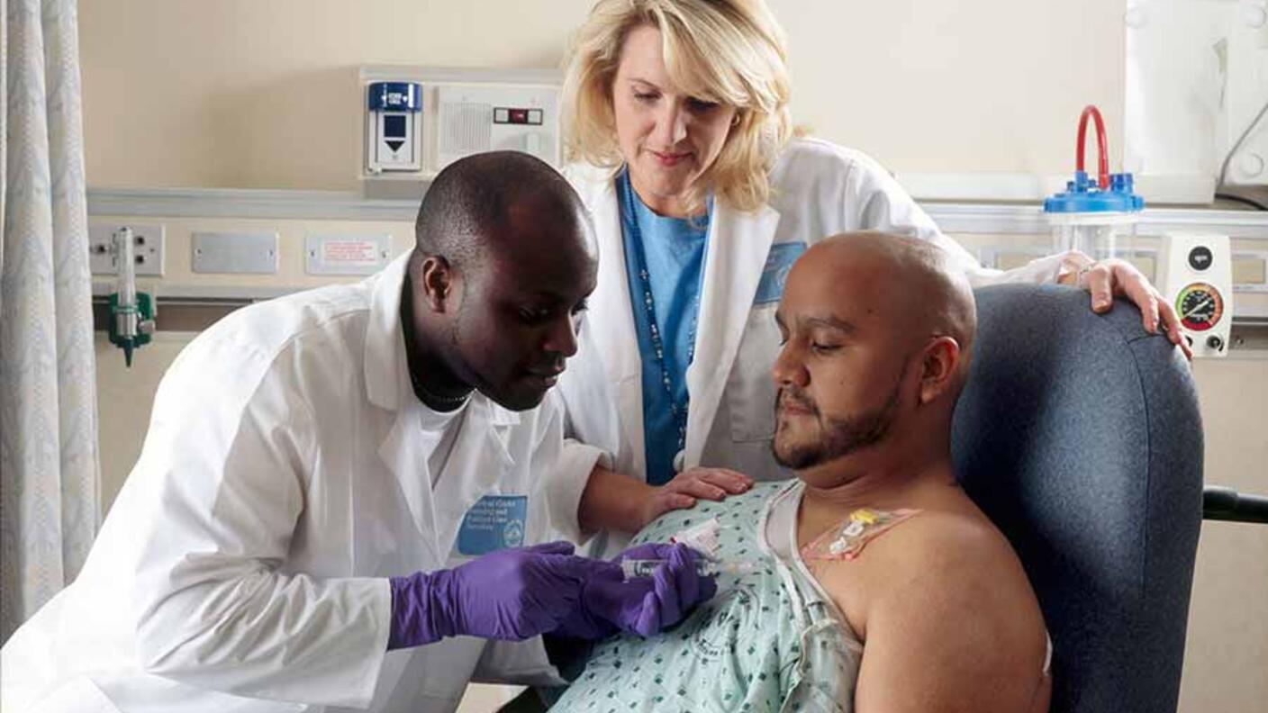 Two medical professionals providing chemotherapy treatment to a patient who has a port in his chest, with one healthcare worker administering the treatment while the other provides support in a hospital setting.