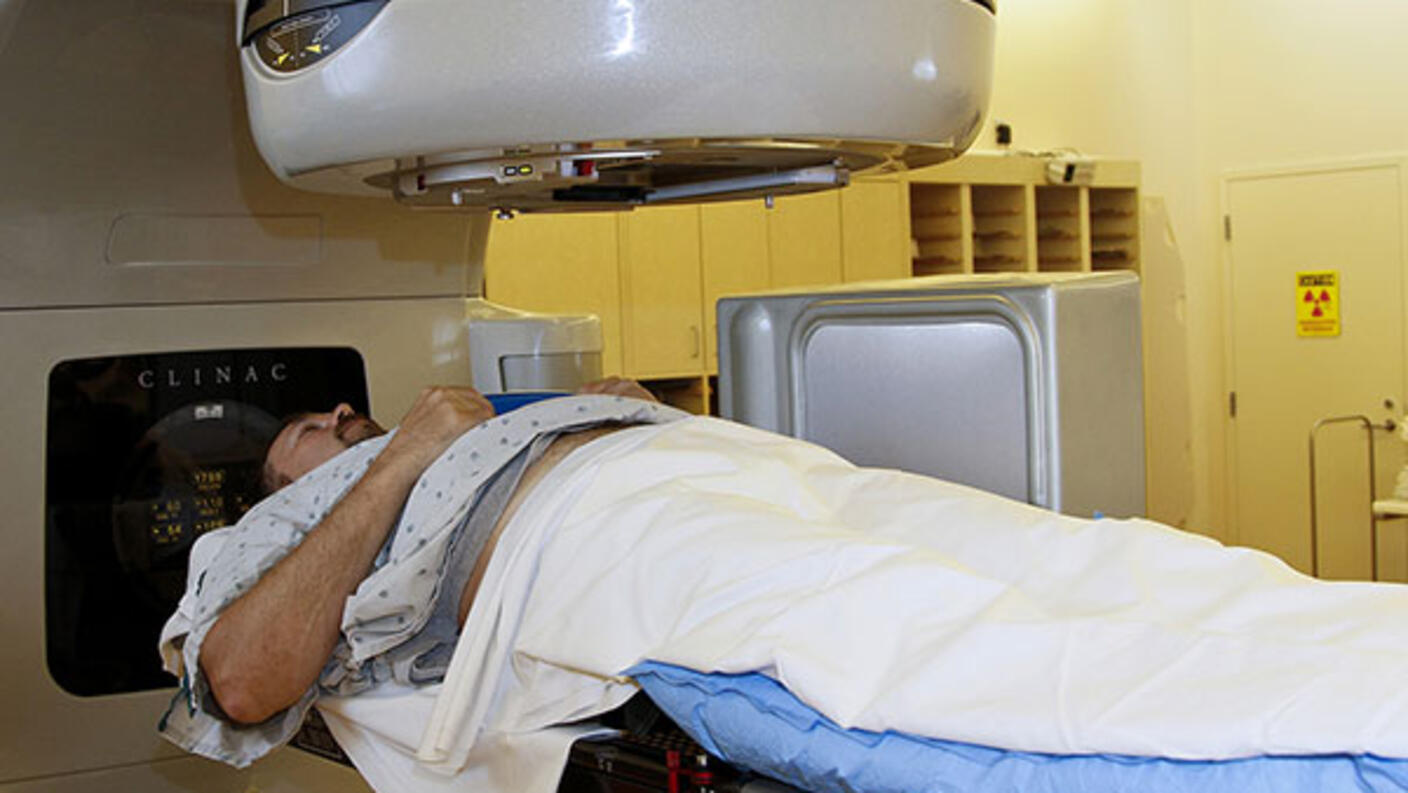 A patient lays on his back under a radiation machine. The patient is dressed in a hospital gown and covered with a blanket. The machine is large and grey with a cylindrical section directly above the patient’s chest. The background is tinted yellow and there are cabinets and a door.