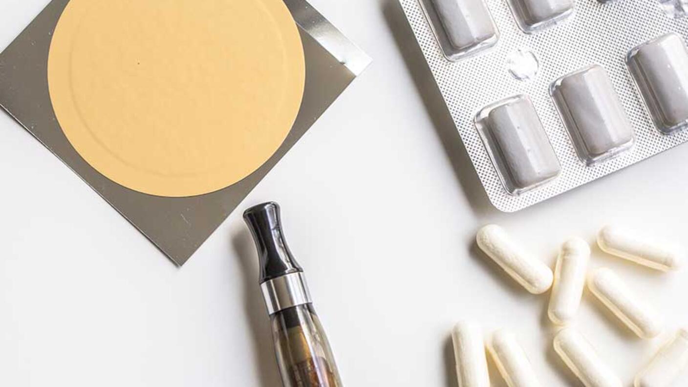 Various smoking cessation products including an e-cigarette device, nicotine patches, pills in blister packs, and white capsules arranged on a white surface.