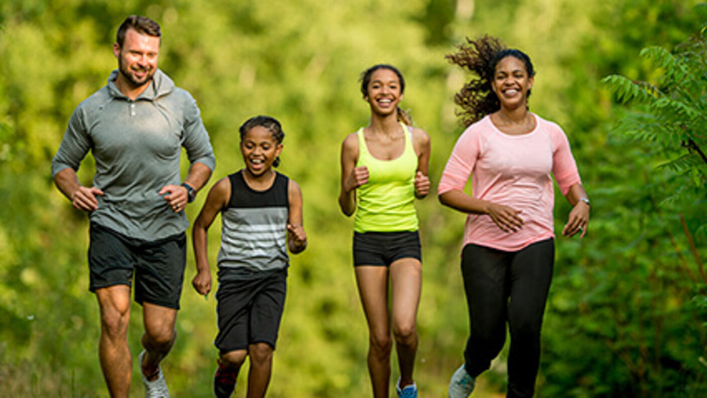 Father, mother, and two daughters run together surrounded by park greenery.