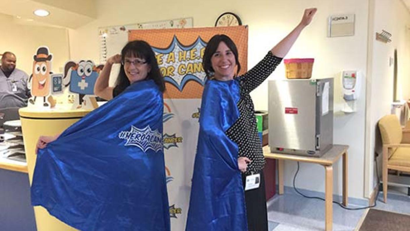 Becky Owens (left) and Sara Rothschild (right) smiling and wearing bright blue superhero capes pose in a hospital environment. The capes feature the words "Be a Hero for Cancer."