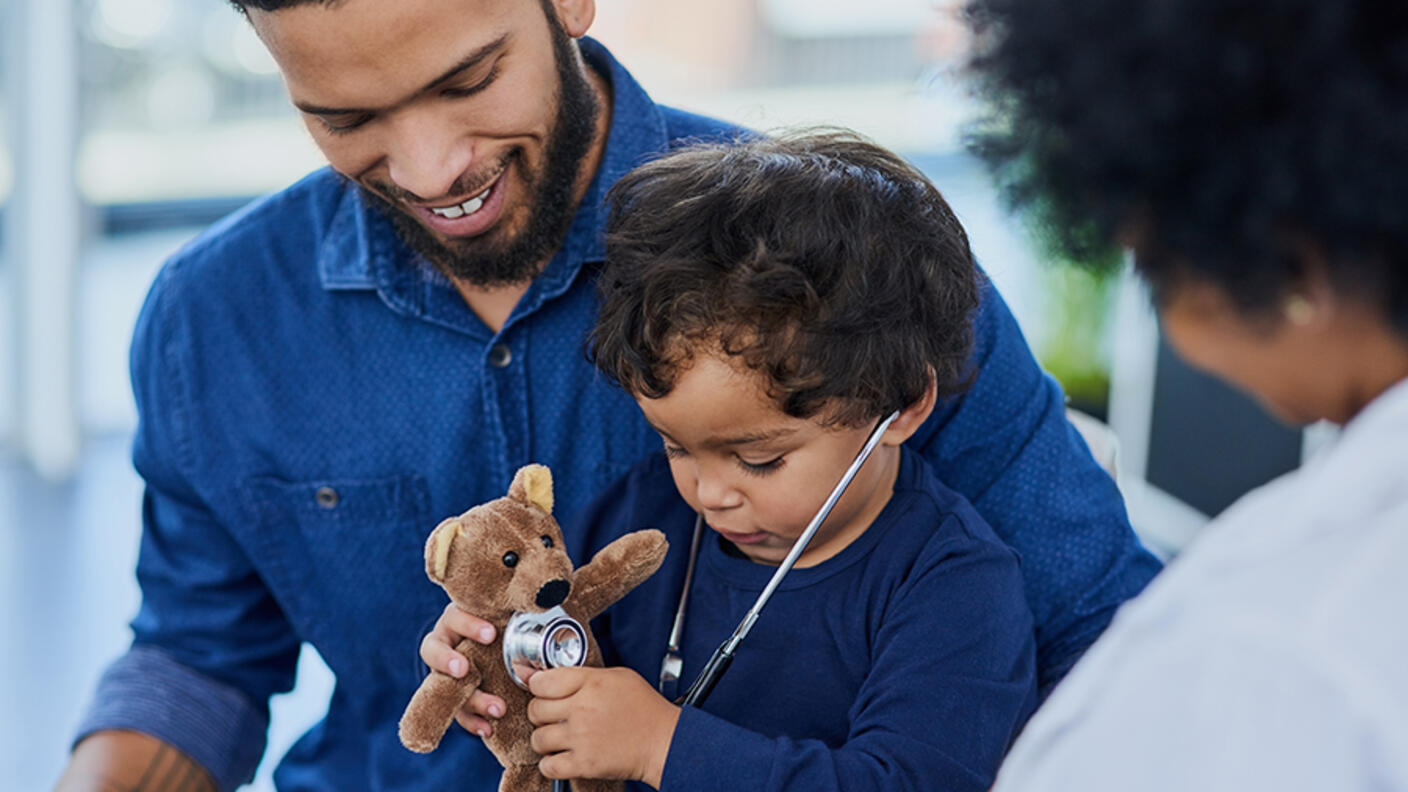 A pediatric patient wears a stethoscope and holds a stuffed bear as his father smiles at him