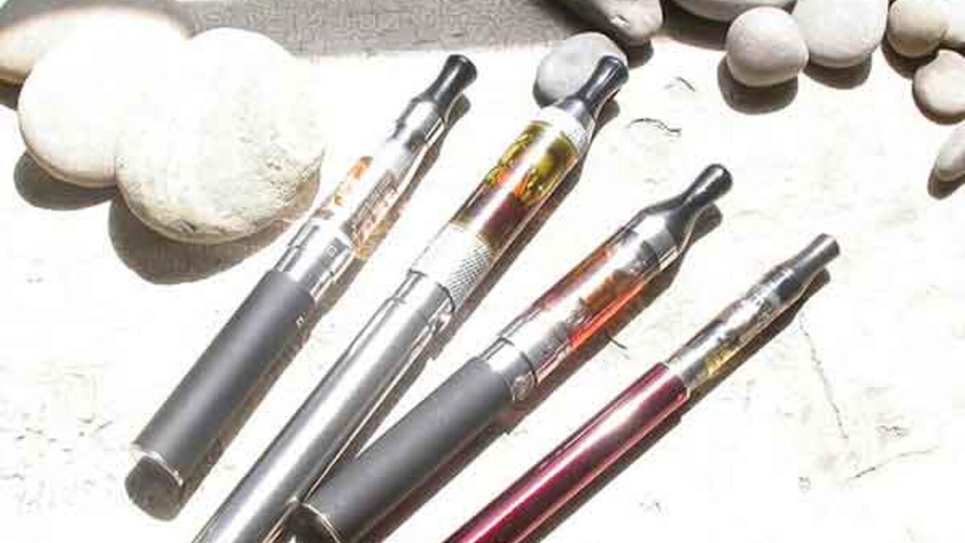 Five e-cigarettes with various colored tanks and cartridges scattered on a light wooden surface.