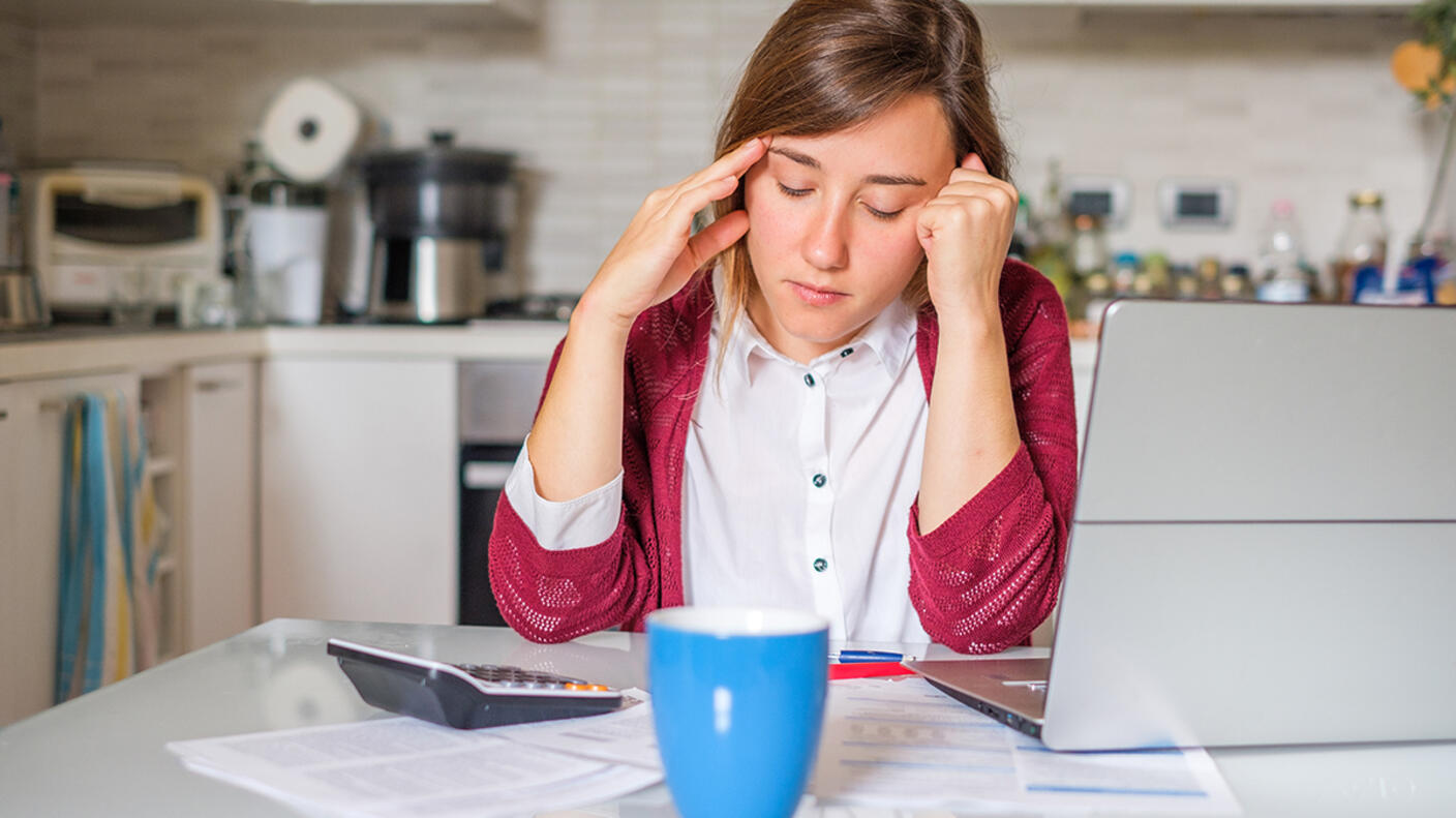Young woman sitting at the kitchen table, looking down at several bills, a calculator, and a laptop.