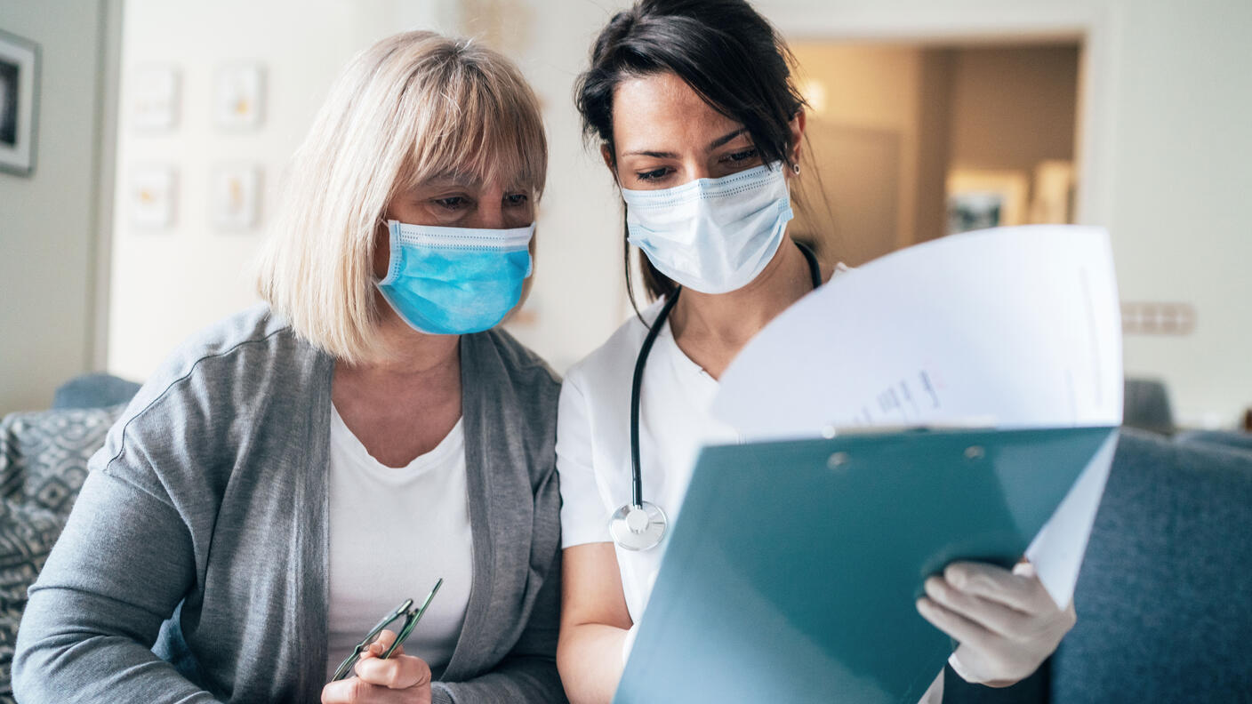 A nurse wearing a face mask talking with an older female patient wearing a face mask.