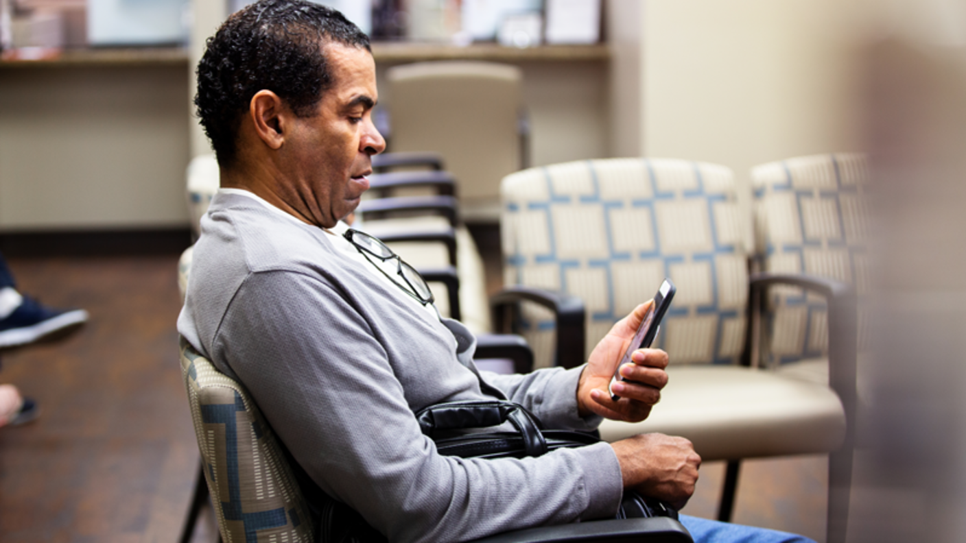 A Hispanic man sitting in a waiting room looking at his smartphone.