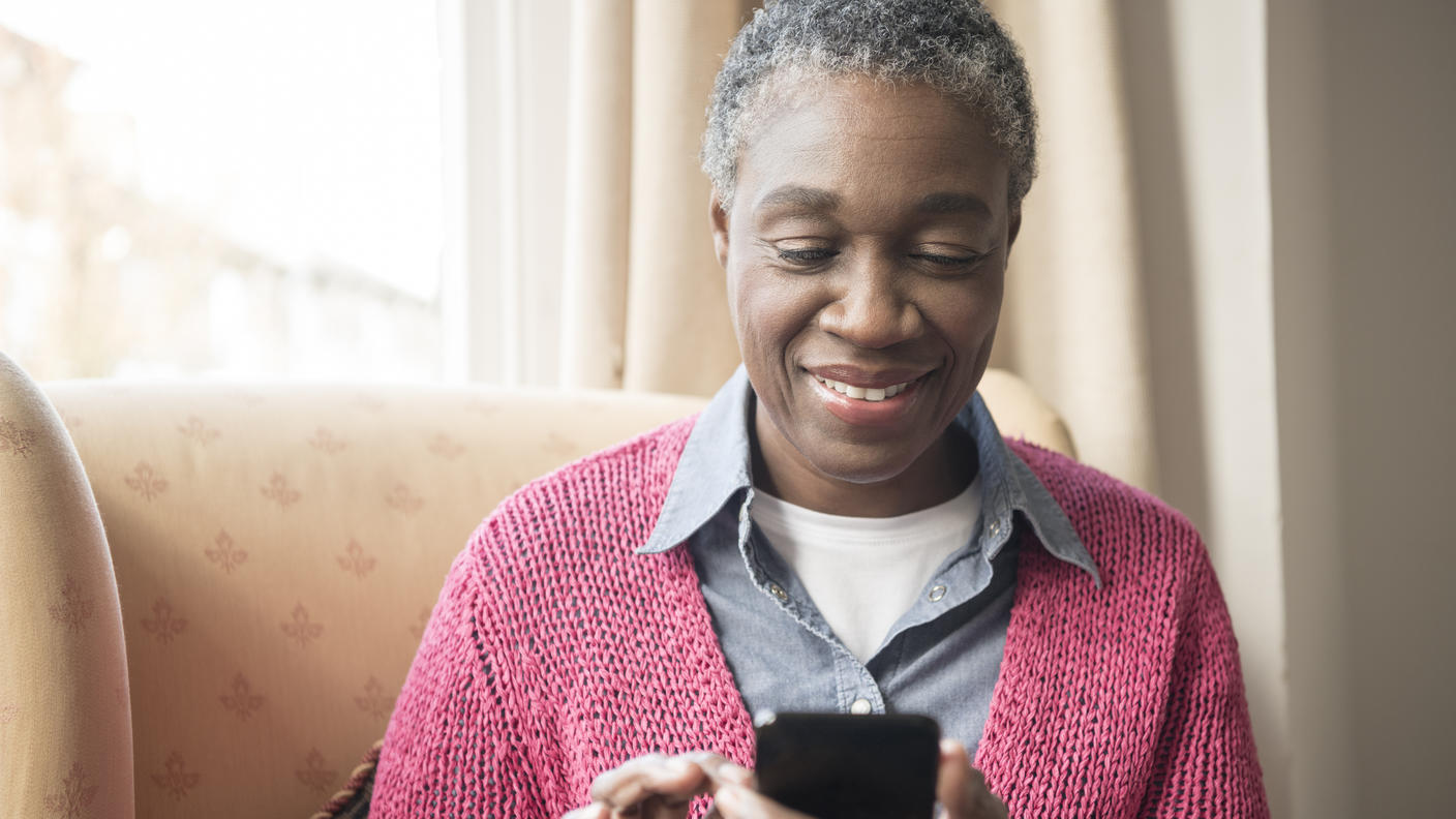 Woman with short hair sitting comfortably in a chair by a window, smiling while using a smartphone. She is wearing a pink cardigan over a denim shirt. 