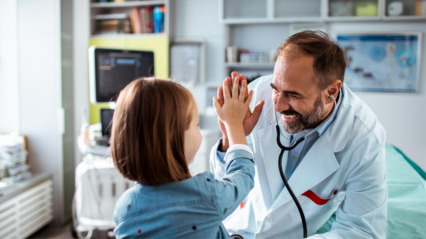 Male doctor giving a young female child a high five in an exam room.