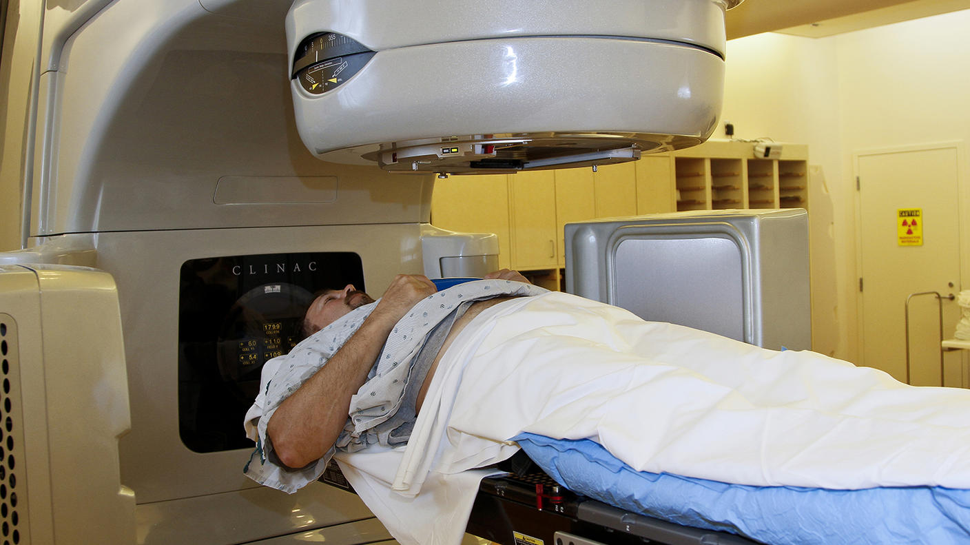 A patient in a hospital gown lies on a treatment table beneath a large linear accelerator machine during a radiation therapy session.