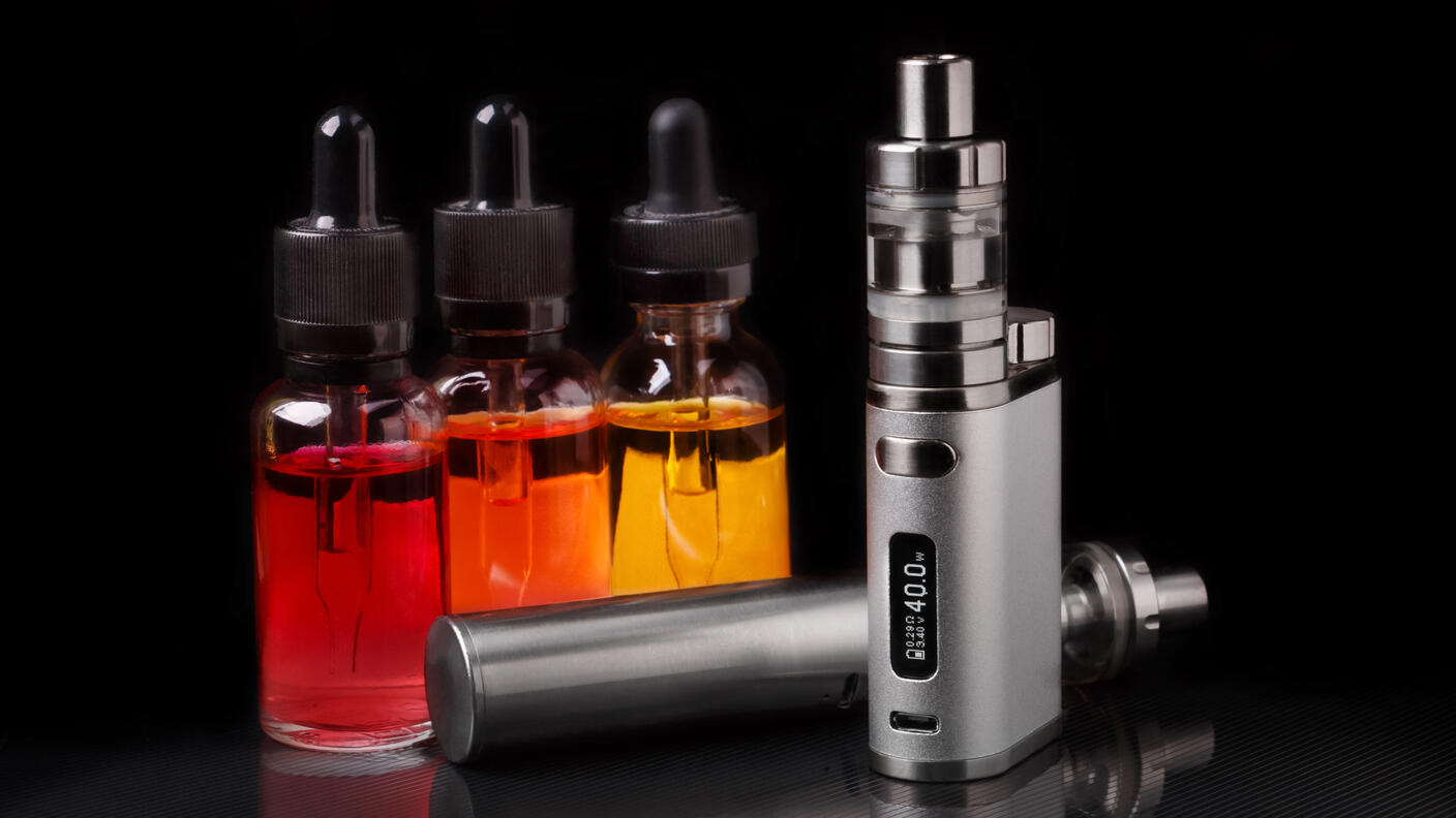 An image of an e-cigarette machine in the foreground with three small bottles that have eyedropper tops in the background. The small bottles contain liquids colored red, orange and yellow; the e-cigarette machine is grey with metal touches.