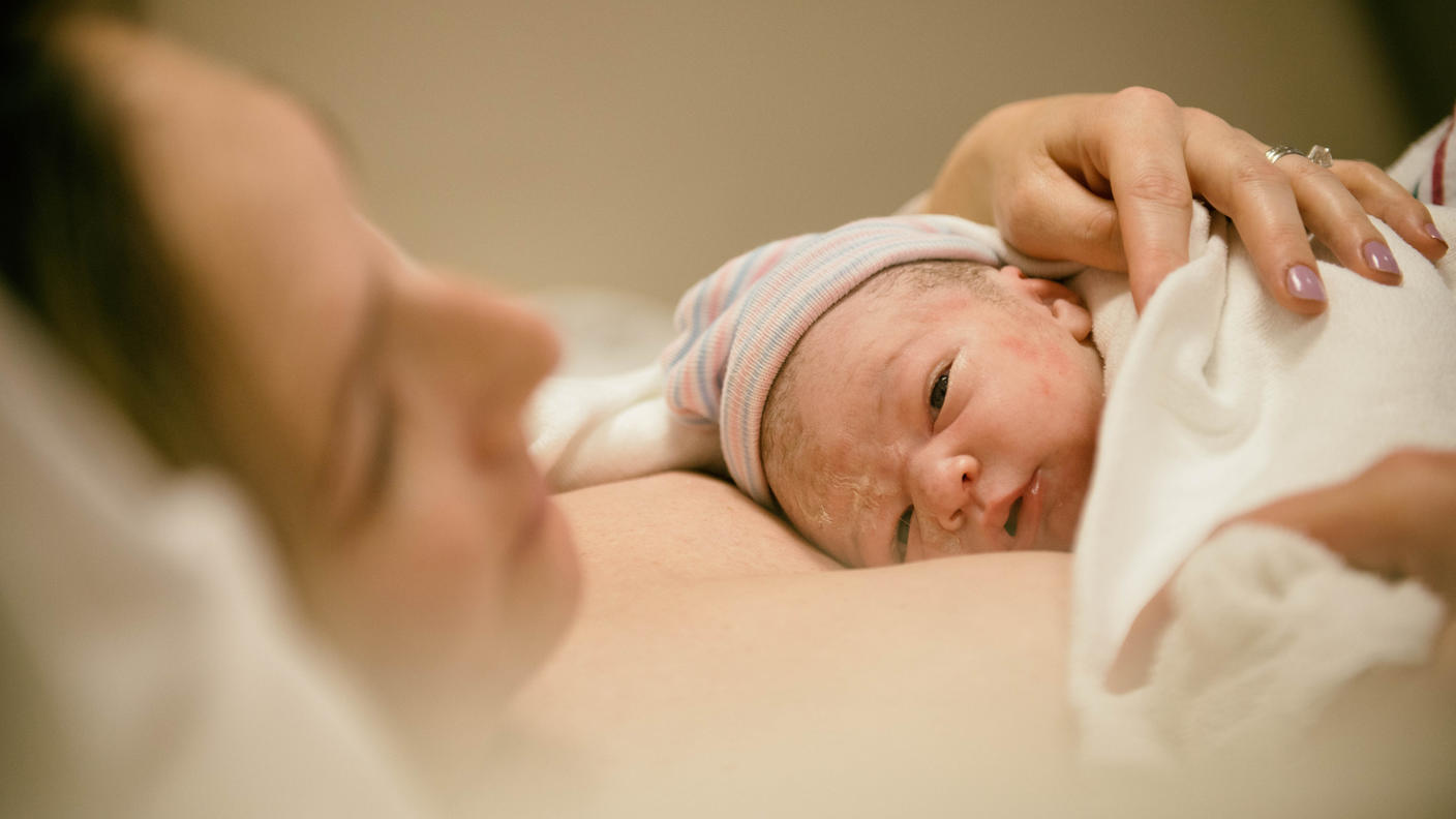A close-up of a mother holding a newborn baby against her chest. The baby has a small hat and is covered with a blanket. The woman’s head rests against a pillow.