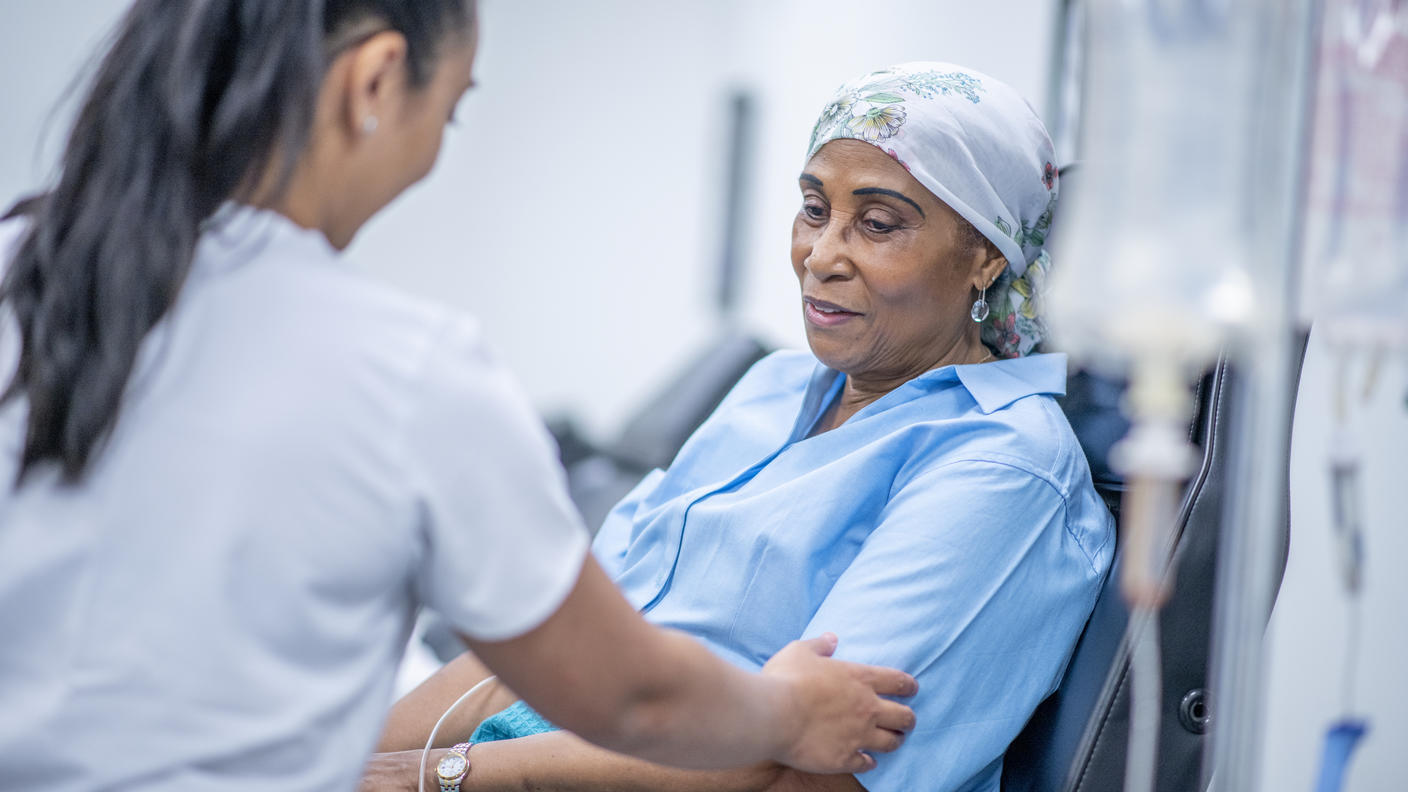 A nurse holds the arm of a sitting patient: an elderly woman with a head covering. A blurry IV bag is in the foreground; the background indicates a treatment center.