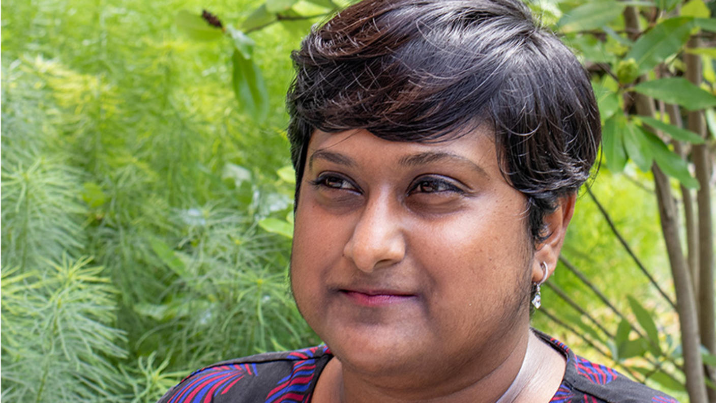 Deepa Prasad stands in front of a green, wooded background while looking at something off camera to her right. She is a medium-skinned woman with short, dark hair and is wearing a dark blue top with a light purple brushstroke print. 