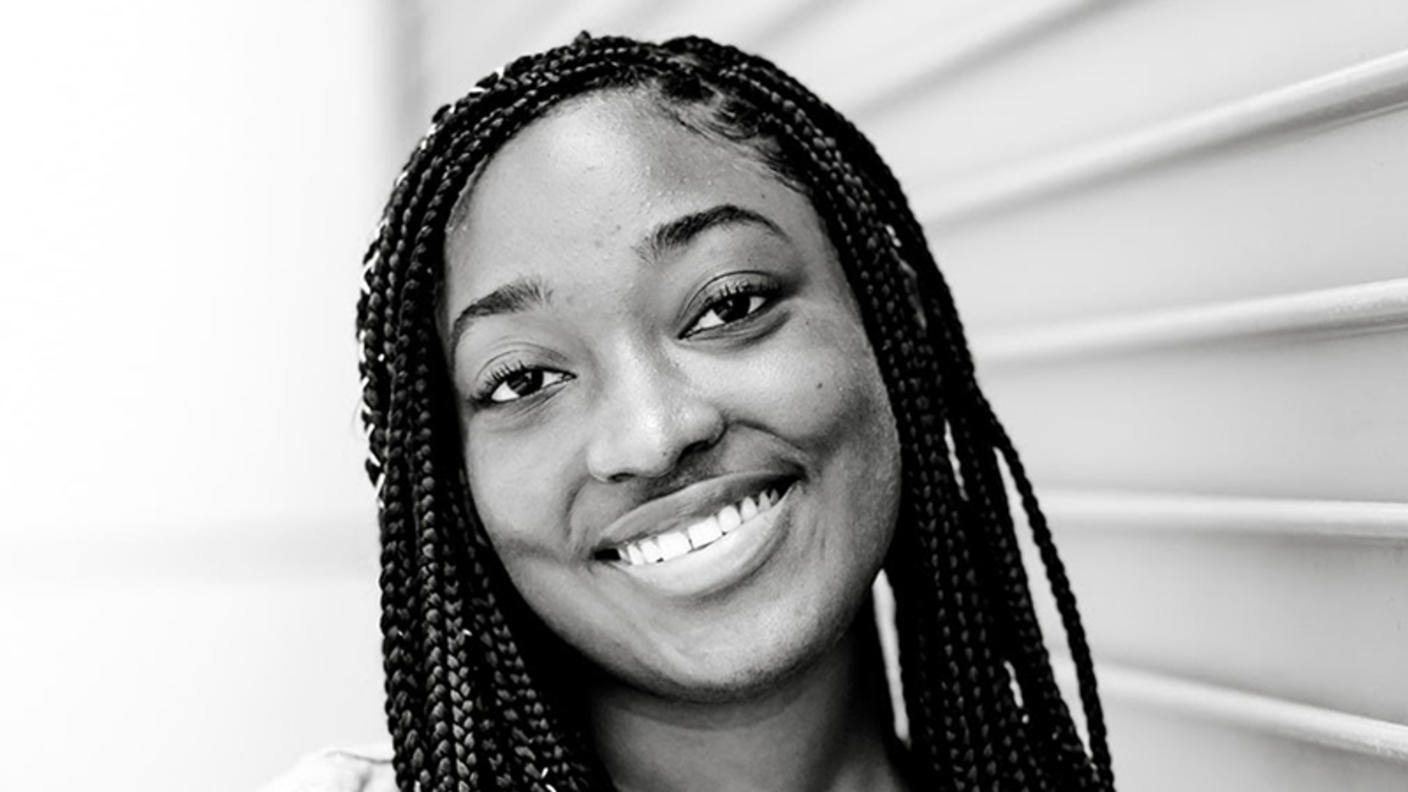 Black and white photo of young, Black woman with braids, standing against a building.