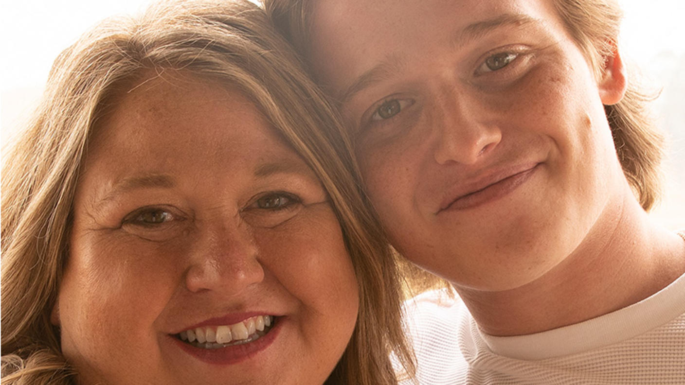 Lisa (a blonde, white woman) stands smiling and leaning into son, Jace (a blonde, white, young adult man).