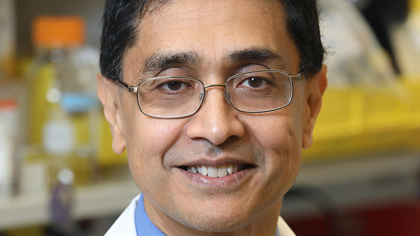 A man with medium brown skin tone and dark hair, wearing glasses, a blue shirt, a navy-blue tie, and a lab coat, is standing in a lab smiling at the camera.
