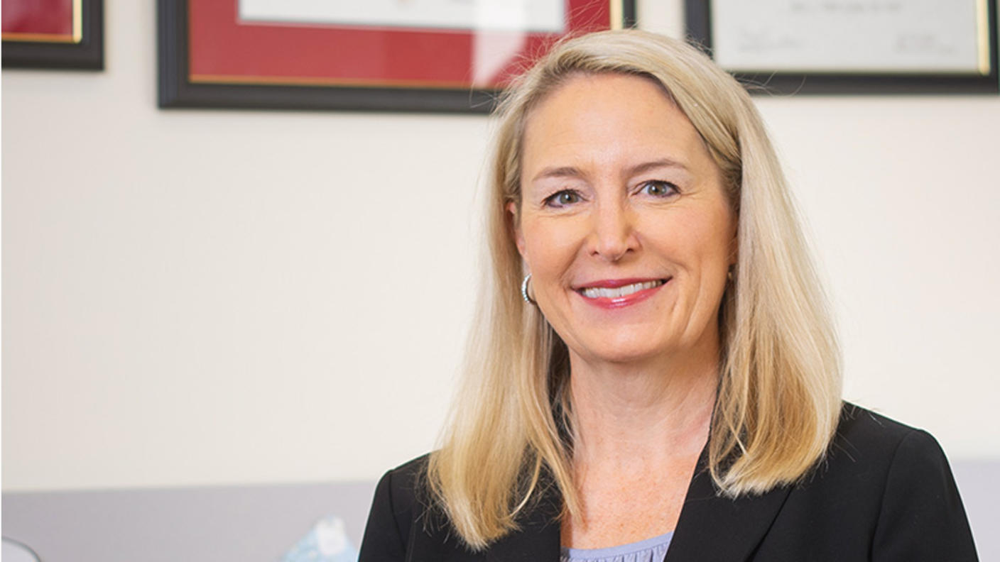 A professional headshot of Dr. Lynne Wagner, a White woman with a light skin tone and blonde hair, wearing a black blazer and blue top while smiling at the camera and standing in her office.